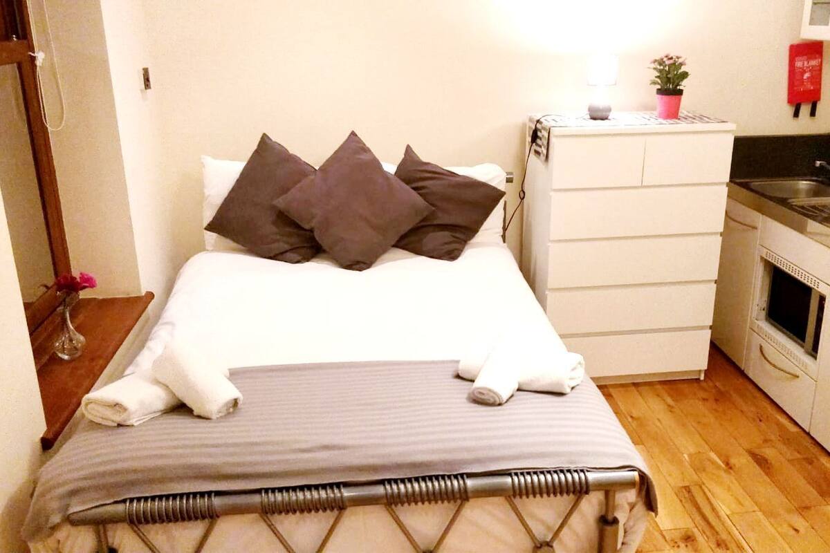 A small, compact bedroom with a bed, a bedside table, a dresser, and a kitchenette.