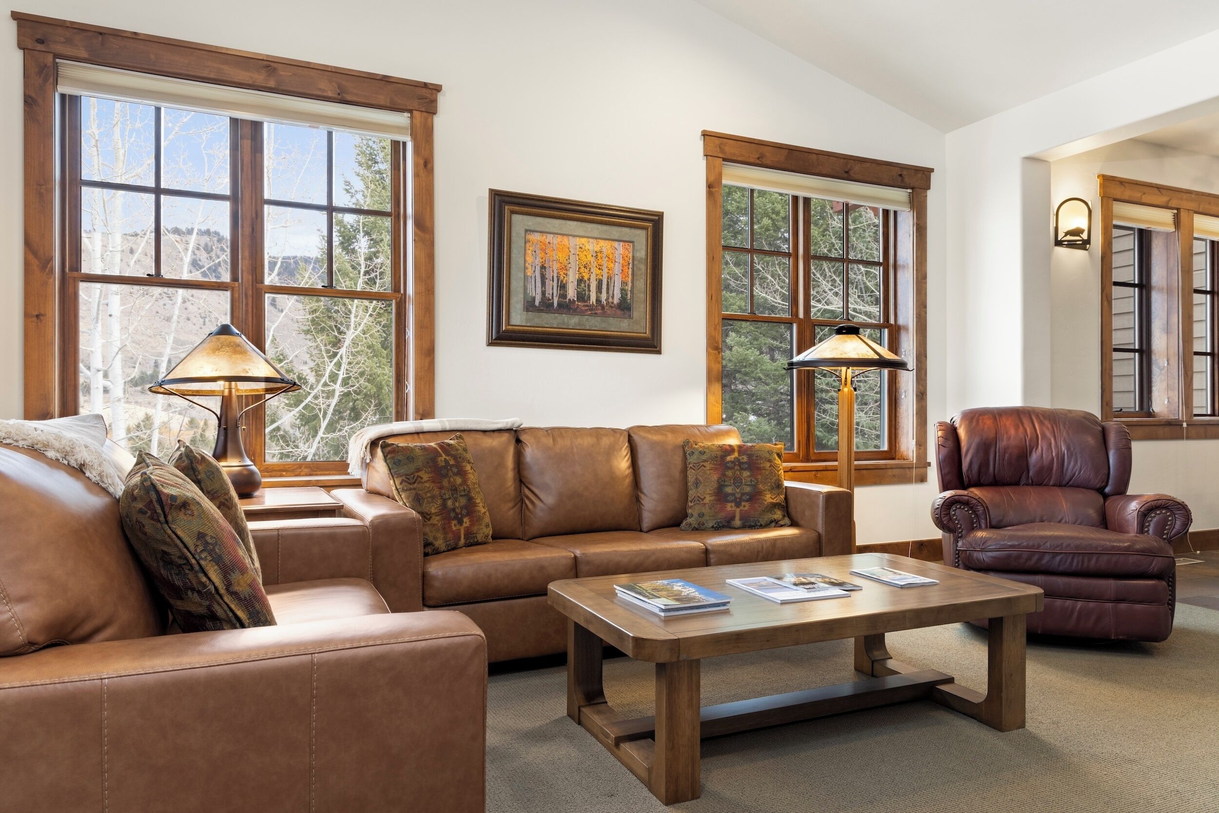 Ein Wohnzimmer mit einem braunen Ledersofa, einem Holztisch und einem Ledersessel. Zwei Fenster mit Holzrahmen und ein Gemälde an der Wand sind zu sehen.