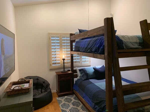 A bunk bed with a wooden frame, a bedside table with a lamp, a bean bag chair, a framed picture, and a window with blinds.