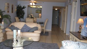 A living room with a beige sofa, a glass coffee table, and a blue rug. There is a ceiling fan, a mirror, and a door leading to another room. - Sand Castle III Luxury Condo, Free nights for select dates. (Indian Shores)