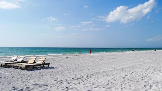 A beach with white sand and a clear blue sky. There are lounge chairs and a person walking in the distance. - Sand Castle III Luxury Condo, Free nights for select dates. (Indian Shores)