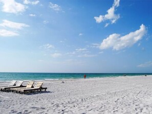 A beach with white sand and a clear blue sky. There are lounge chairs and a person walking in the distance.
