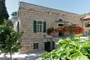 A stone building with green shutters, a balcony with red flowers, and a small door.