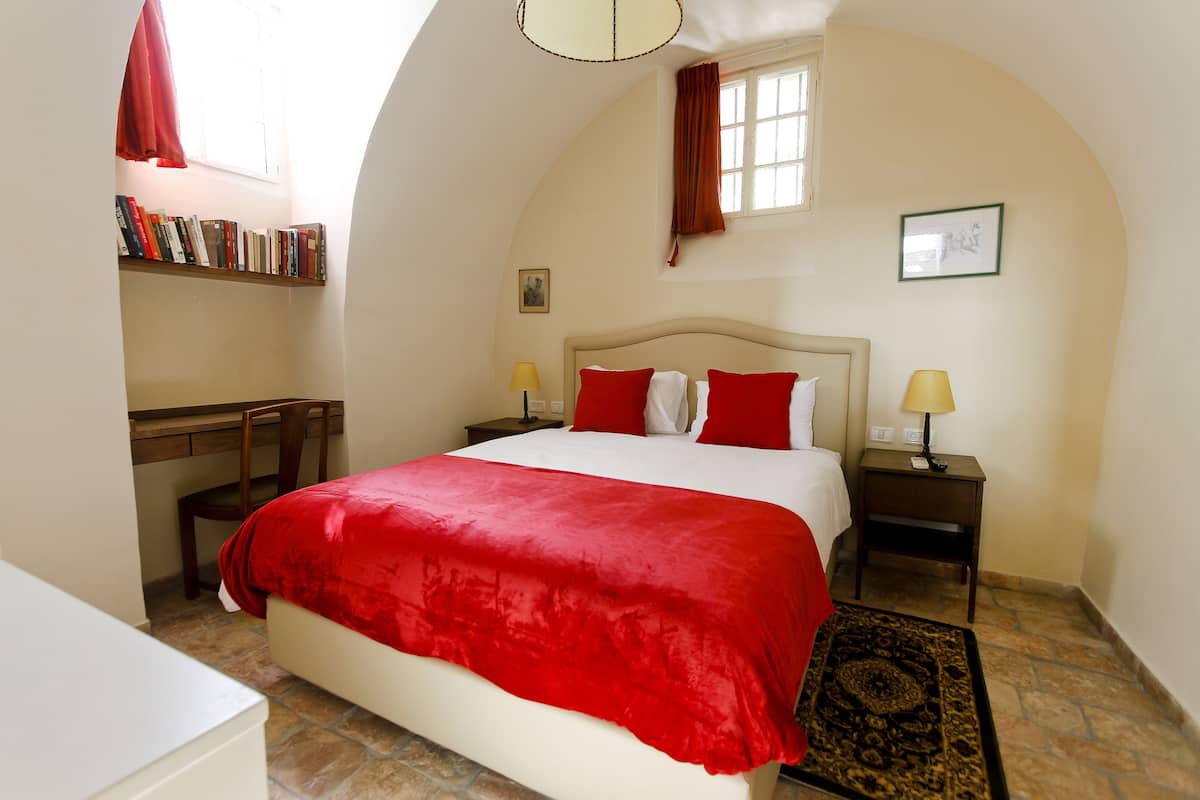 A bedroom with a large bed, two bedside tables with lamps, a desk, a chair, a bookshelf, and a window with curtains.