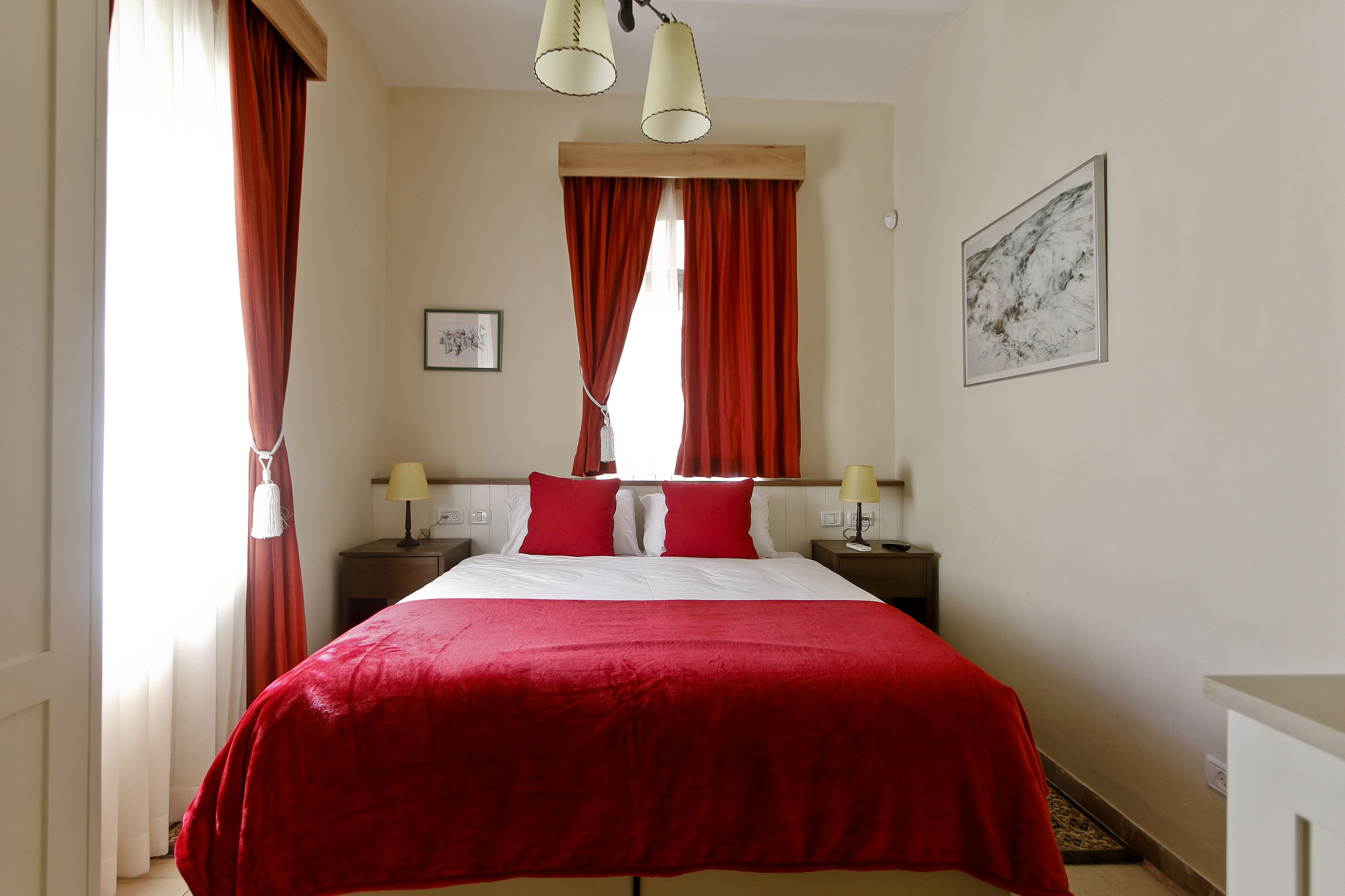 Habitación de hotel con una cama grande, cortinas rojas, dos lámparas de noche, un cuadro enmarcado en la pared y una mesita.