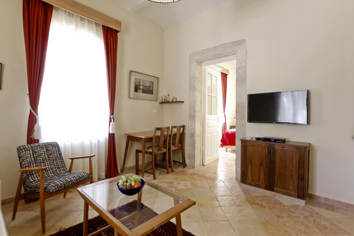 A living room with a sofa, a glass coffee table with fruit, a TV on the wall, a wooden cabinet, and a dining area with chairs and a table.