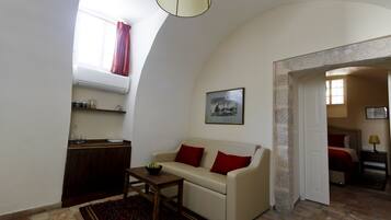 A living room with a sofa, a table, a window with curtains, a framed picture on the wall, and a doorway leading to another room.