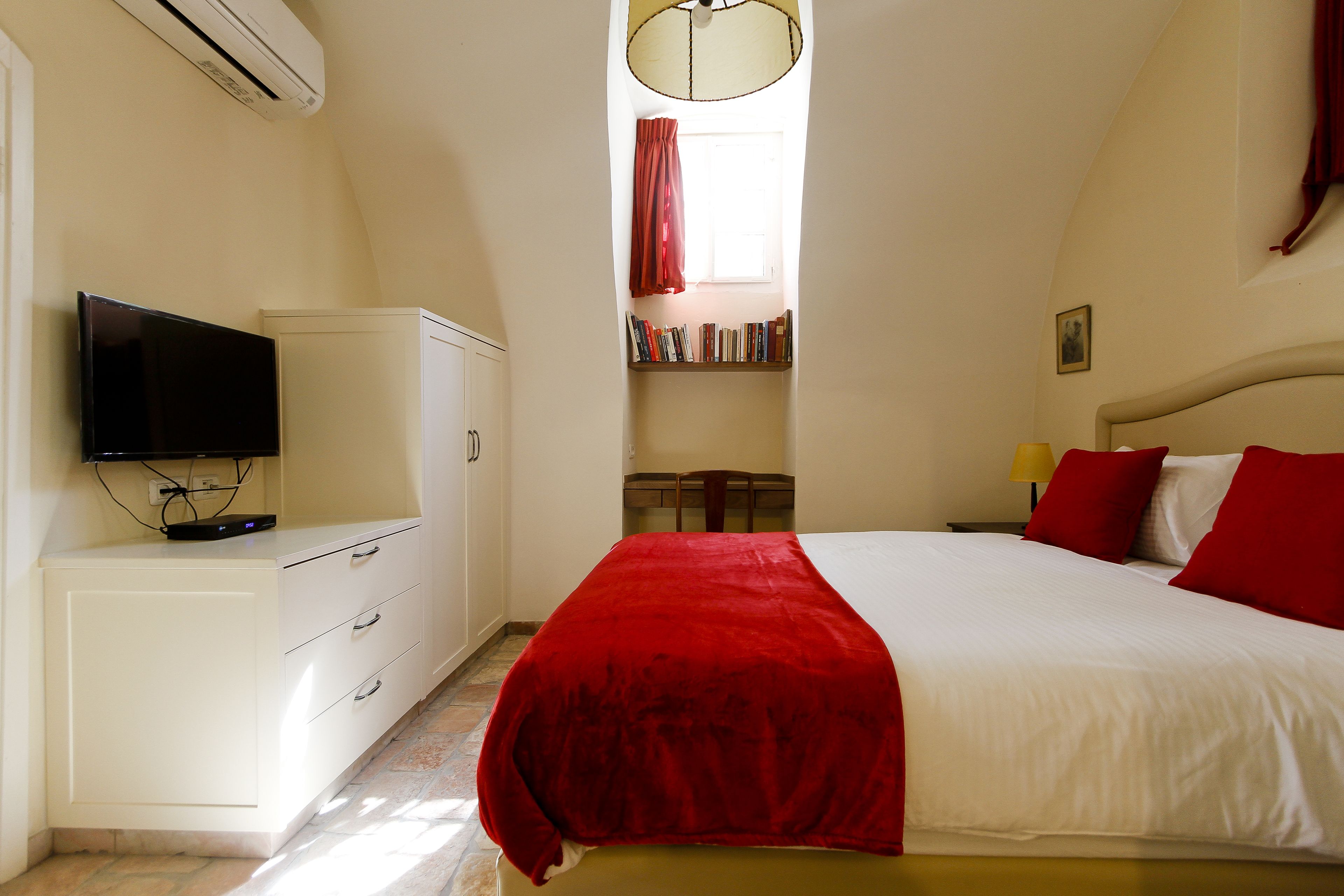 Une chambre à coucher avec un lit, un téléviseur posé sur un meuble, une bibliothèque et une fenêtre avec des rideaux.