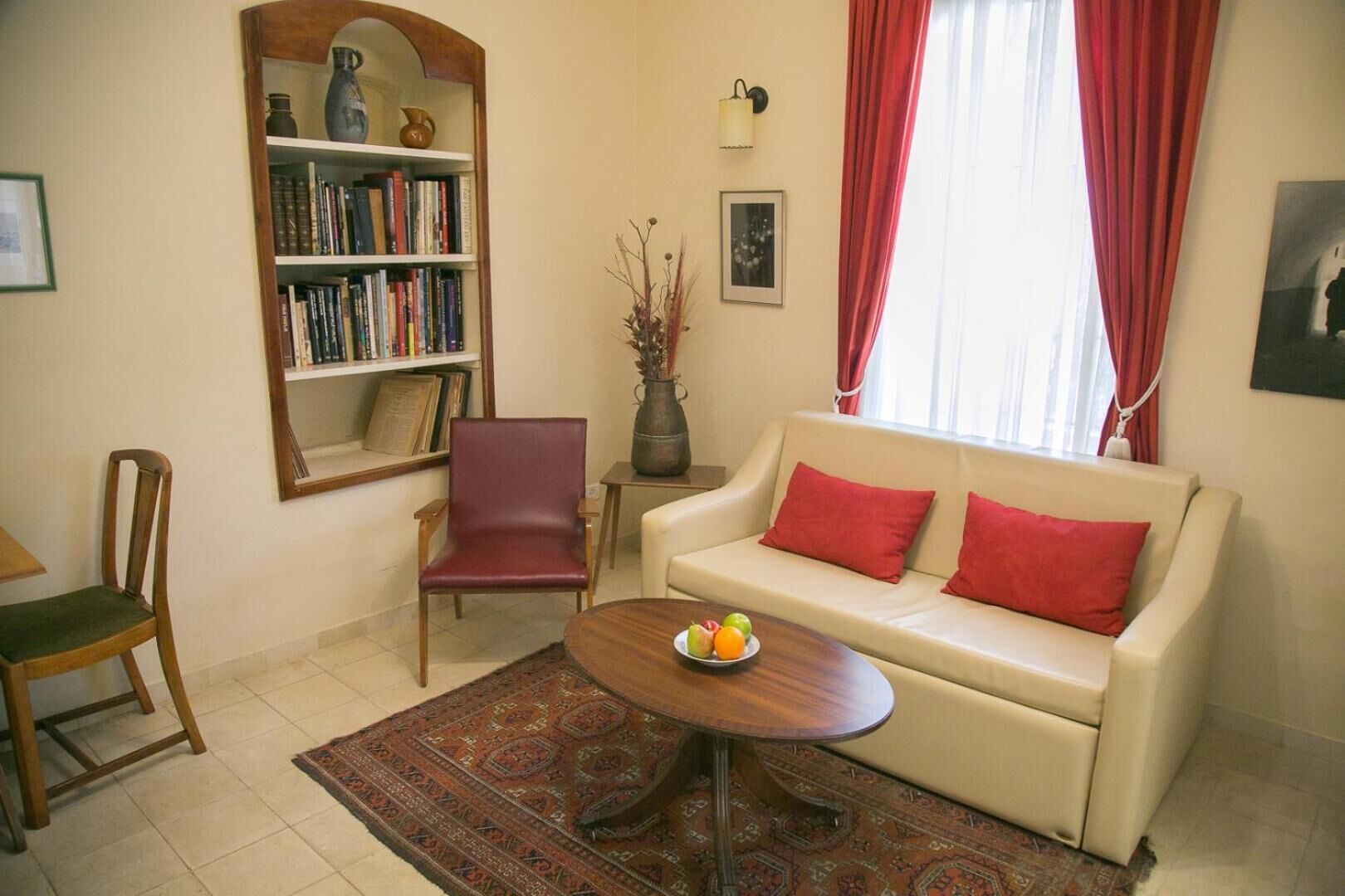 Un salón con un sofá color crema, cojines rojos, una silla de madera, una estantería, una mesita con fruta y una alfombra estampada.
