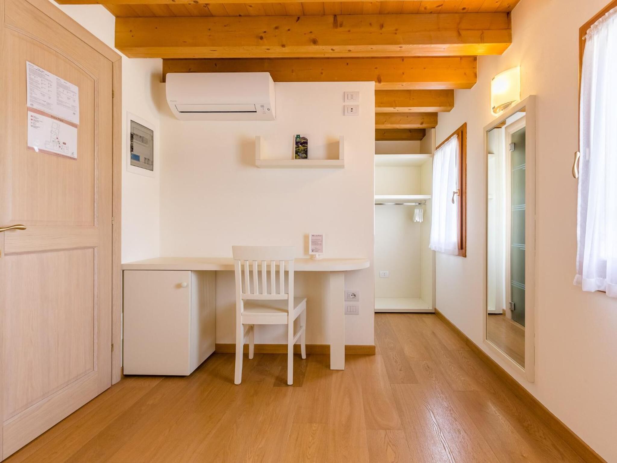 Quarto compacto com teto de madeira, uma pequena escrivaninha branca com cadeira, guarda-roupa branco e ar condicionado branco.