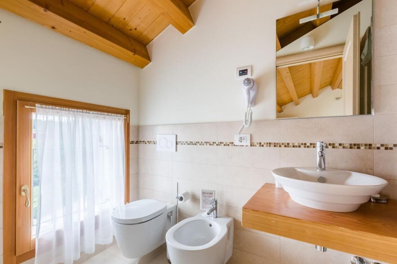 Une salle de bain comprenant des toilettes, un bidet et un lavabo, avec des poutres en bois au plafond et une fenêtre avec des rideaux.