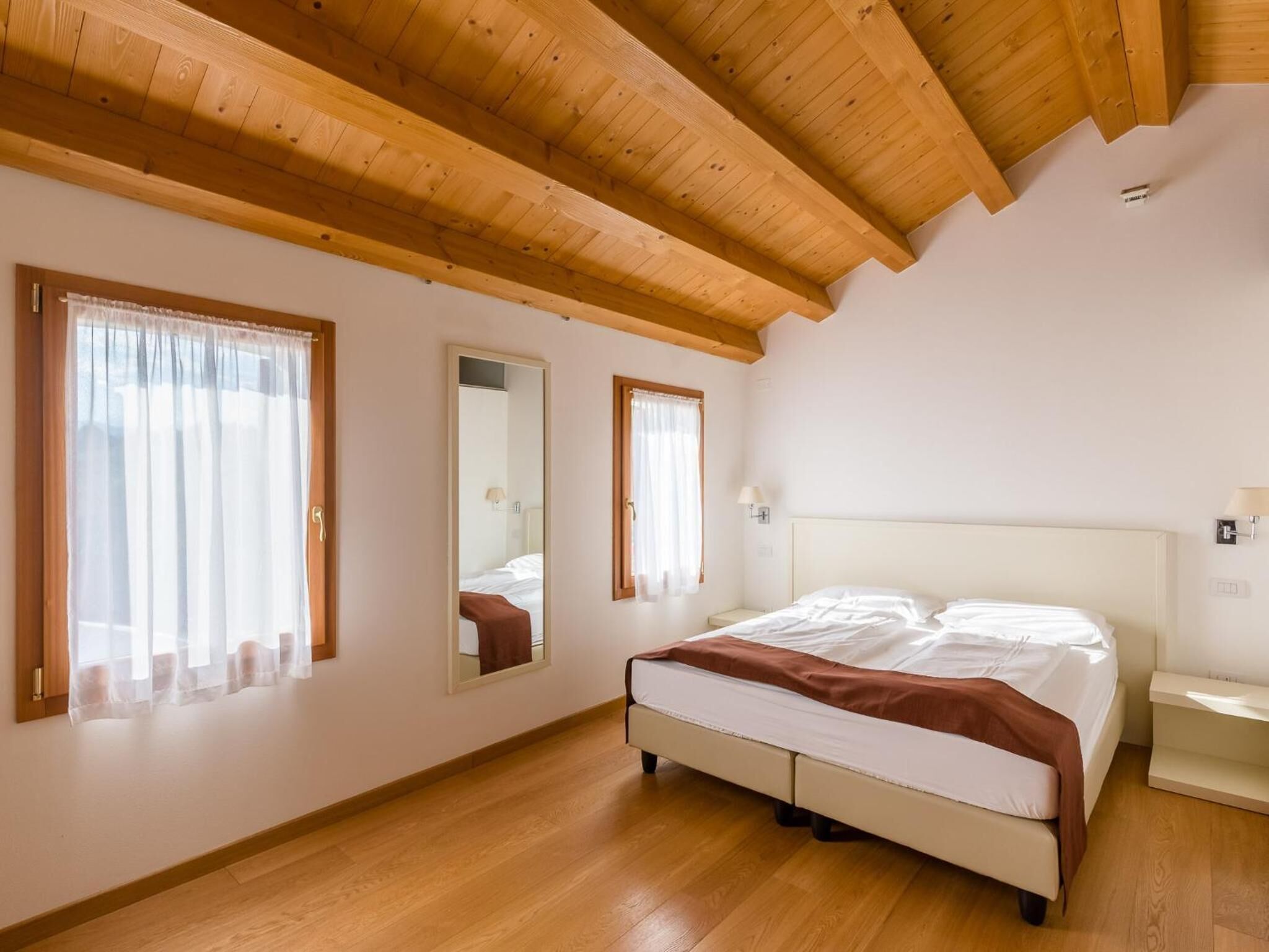 Ein Schlafzimmer mit Holzbalken, einem Bett, einem Nachttisch und zwei Fenstern mit transparenten Vorhängen.