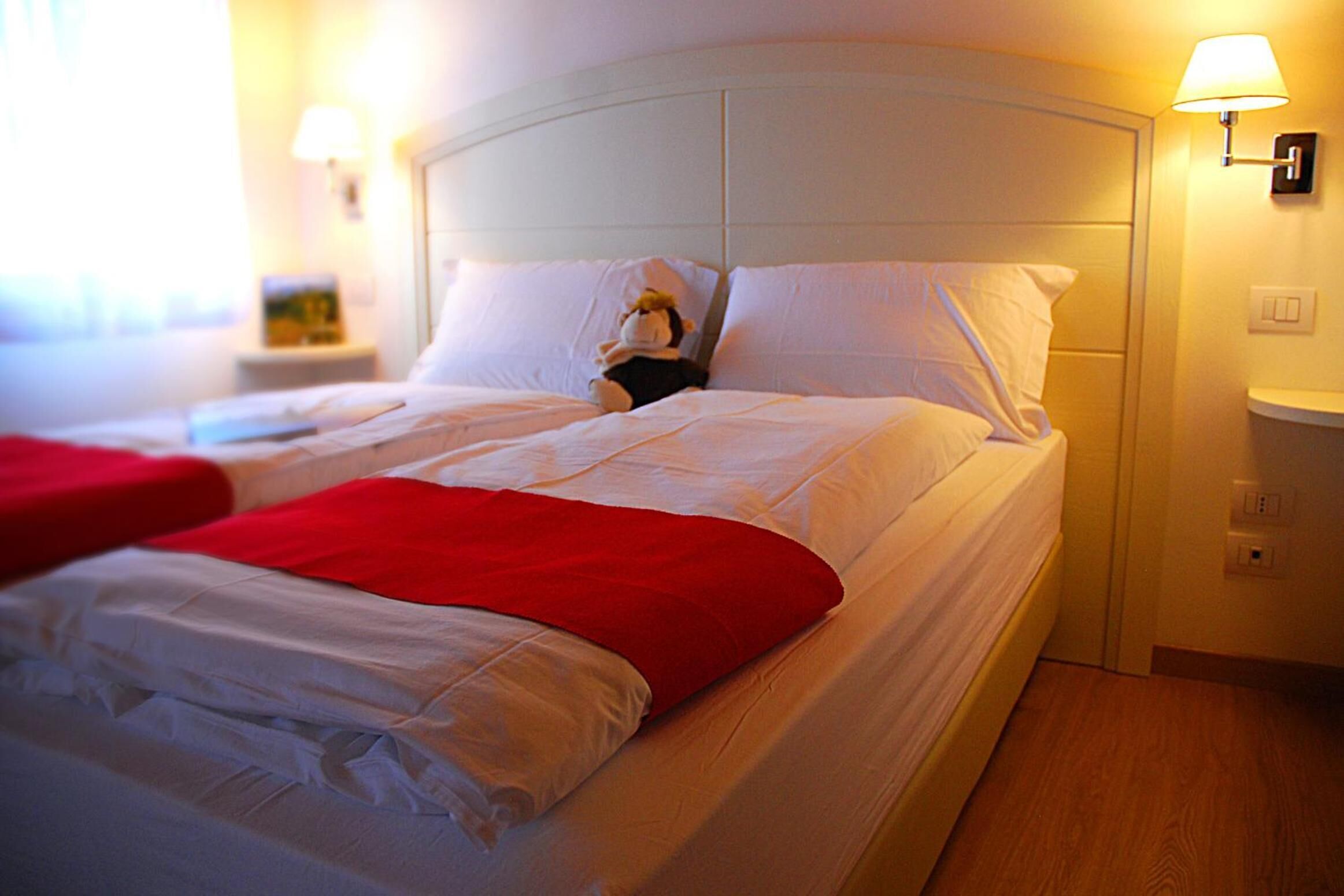 A hotel room with two beds, white bedding, a red blanket on one bed, and a nightstand with a lamp.