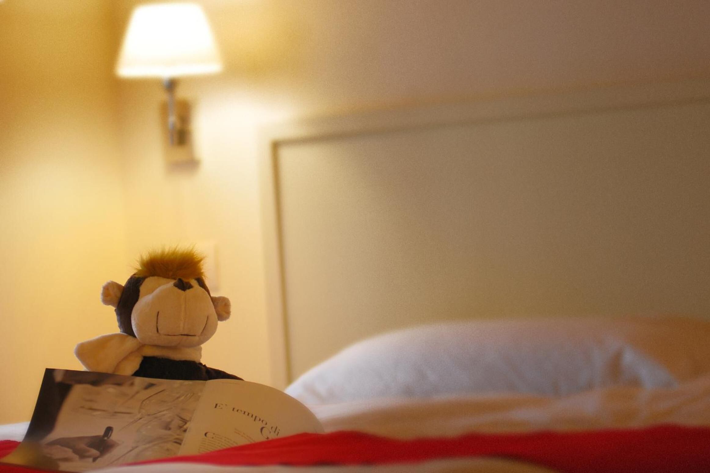 A plush toy reading a book on a bed with white pillows and a red blanket.