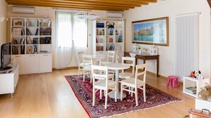 Ein gemütliches Wohnzimmer mit Holzboden, einem Bücherregal, einem Esstisch mit Stühlen, einem rosa Hocker und einer Heizung.