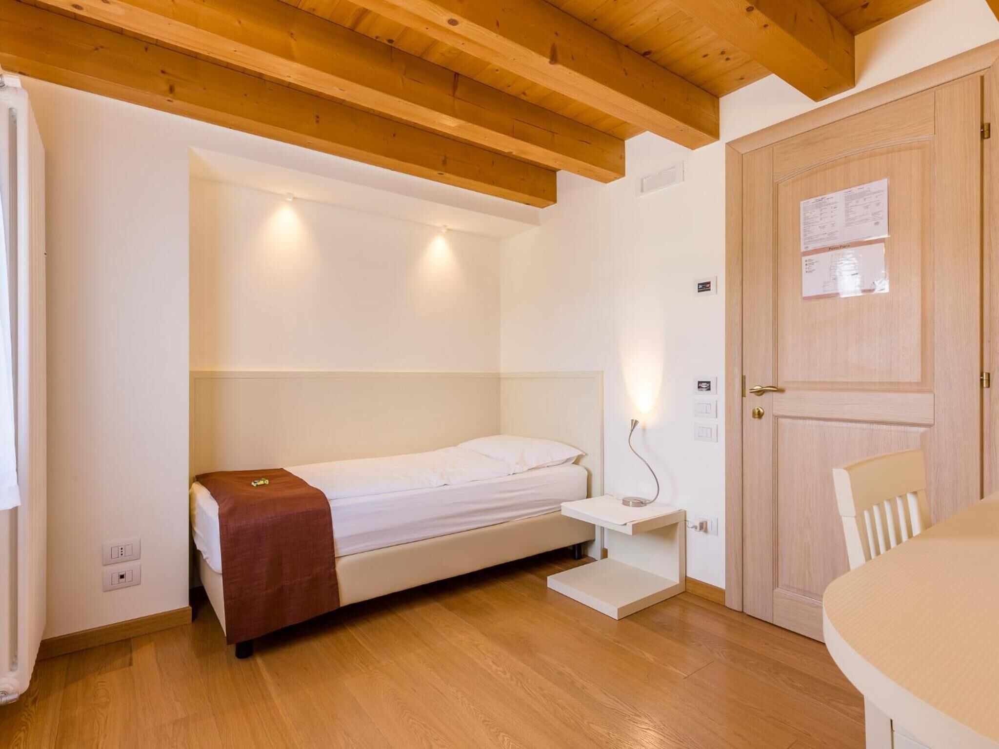 Une petite chambre bien éclairée, avec un lit simple, une table de chevet, un plafond en bois et une porte sur laquelle est affichée une note.