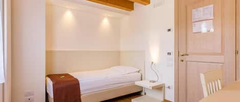 Une petite chambre bien éclairée, avec un lit simple, une table de chevet, un plafond en bois et une porte sur laquelle est affichée une note.