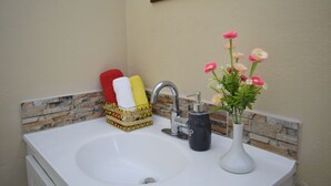 A bathroom sink with a stone backsplash, a mirror above it, and a vase with flowers. - Justinn Apartments (Gros Islet)
