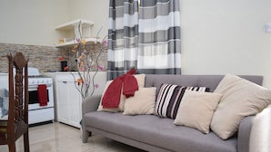 A living room with a grey sofa, a wooden chair, a white stove, and a kitchen area with a brick backsplash. - Justinn Apartments (Gros Islet)