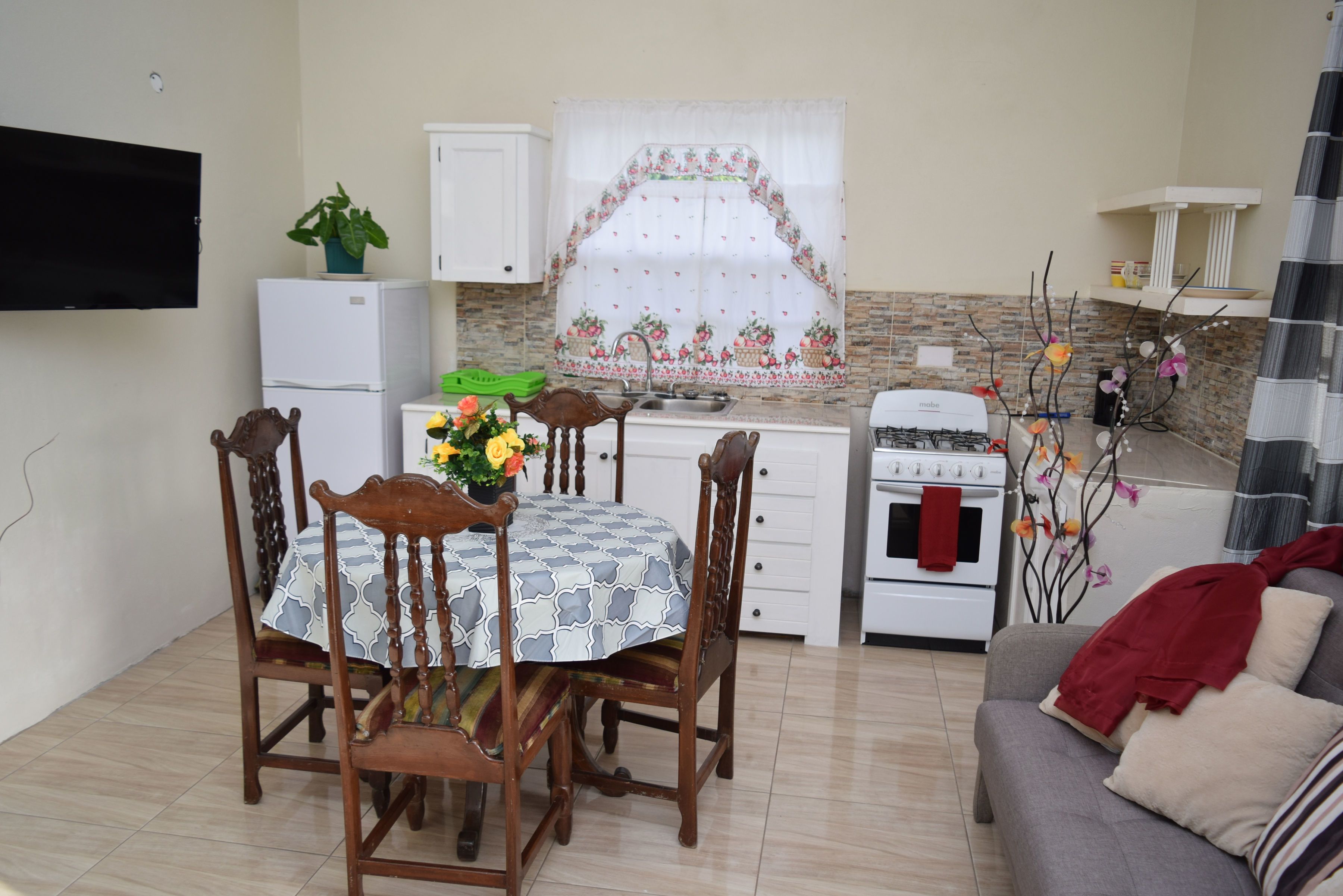 A compact kitchen with a dining table, chairs, a refrigerator, an oven, and a TV mounted on the wall.