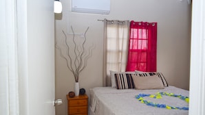 A bedroom with a bed, a nightstand, a vase with branches, and a window with curtains. - Justinn Apartments (Gros Islet)