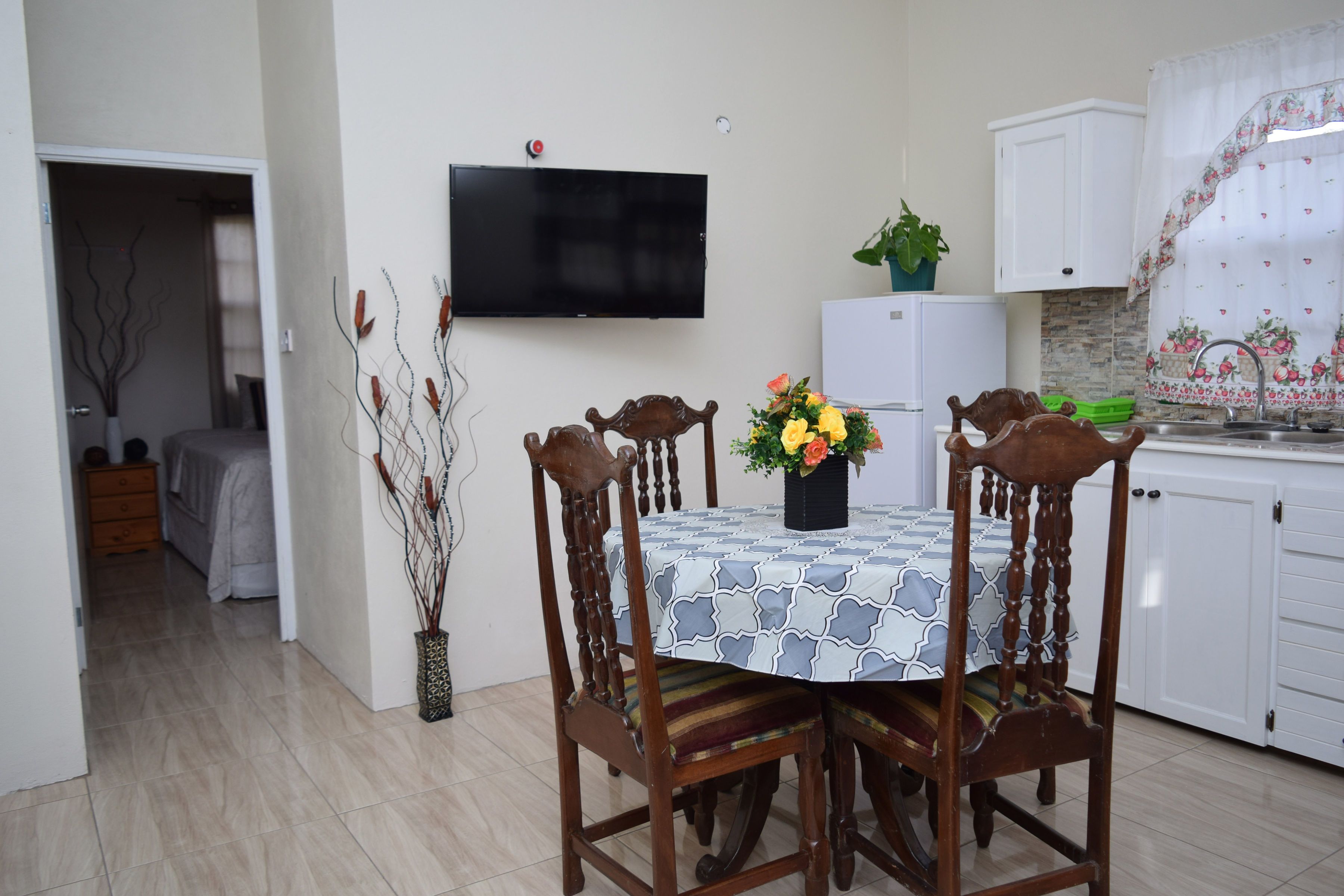 A dining area with a table set for four, surrounded by wooden chairs. A flat-screen TV is mounted on the wall above the table.