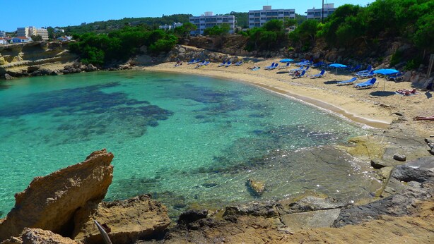 A beach with clear turquoise water, rocky shore, and blue umbrellas. - Proteas Mare Suites (Paralimni)