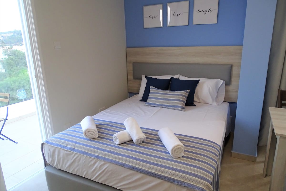 A neatly made bed with striped bedding and rolled towels, a wooden headboard, and a view of greenery outside.