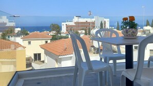 A balcony with white plastic chairs and a table, overlooking a coastal cityscape with buildings and the sea. - Proteas Mare Suites (Paralimni)