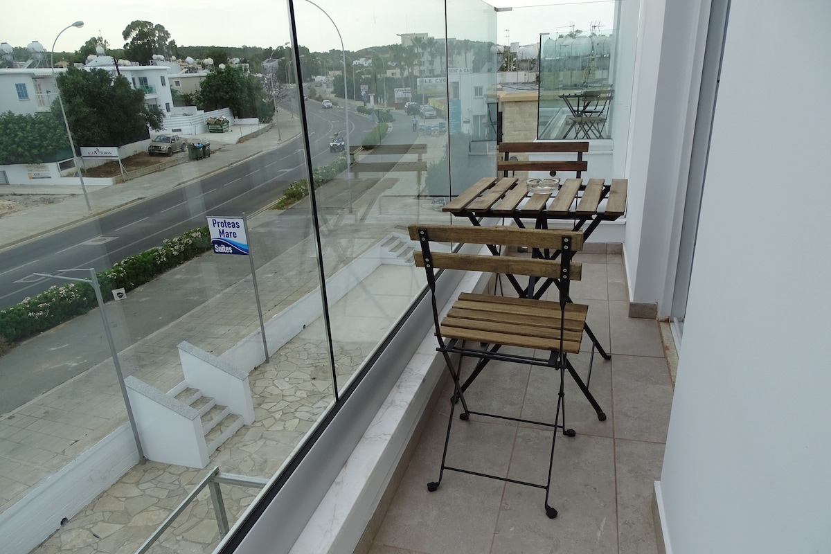 A balcony with a wooden table and chairs, a glass railing, and a view of a street with buildings and a sign for "Proteas Mare Suites".