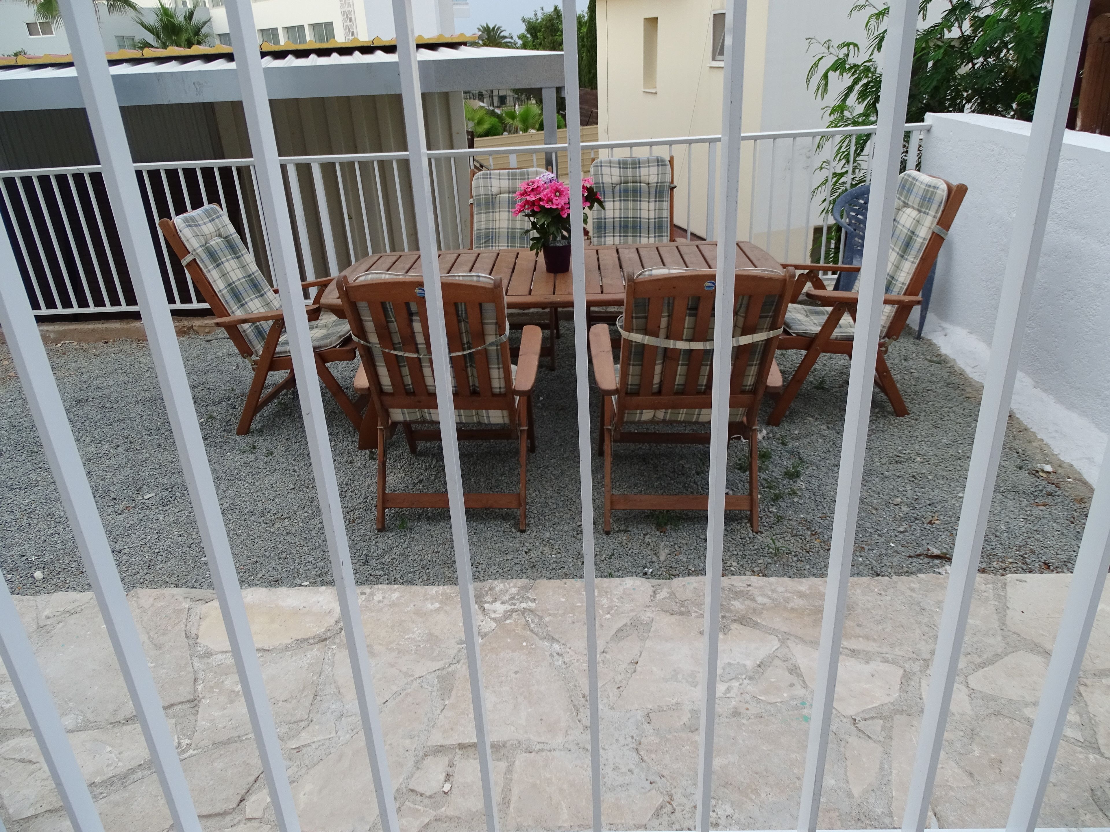 Un patio al aire libre con una mesa de madera redonda y seis sillas, rodeado por una valla de metal blanco.