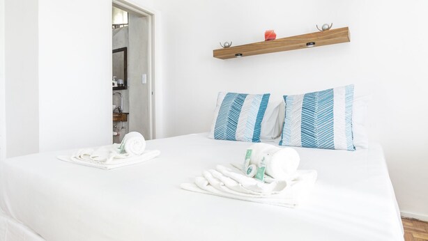 Ein Hotelzimmer mit Bett, zwei Kissen, einer Holzablage mit einem roten Gegenstand und einem durchgegangenen Türrahmen sichtbaren Badezimmer.