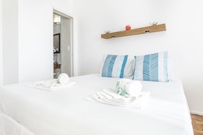 A hotel room with a bed, two pillows, a wooden shelf with a red object, and a bathroom visible through an open door.