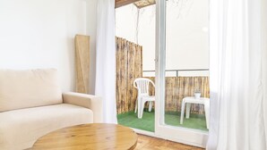 Ein Zimmer mit einem beigefarbenen Sofa, einem Holztisch und einer Schiebetür zu einer Terrasse mit einem Bambuszaun und zwei weißen Plastikstühlen.