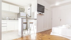 Ein modernes Hotelzimmer mit Holzboden, einem runden Holztisch, einem Bett mit weißer Bettwäsche, einem an der Wand montierten Fernseher und einer Küchenzeile mit weißen Schränken und einem Kühlschrank mit Muster.