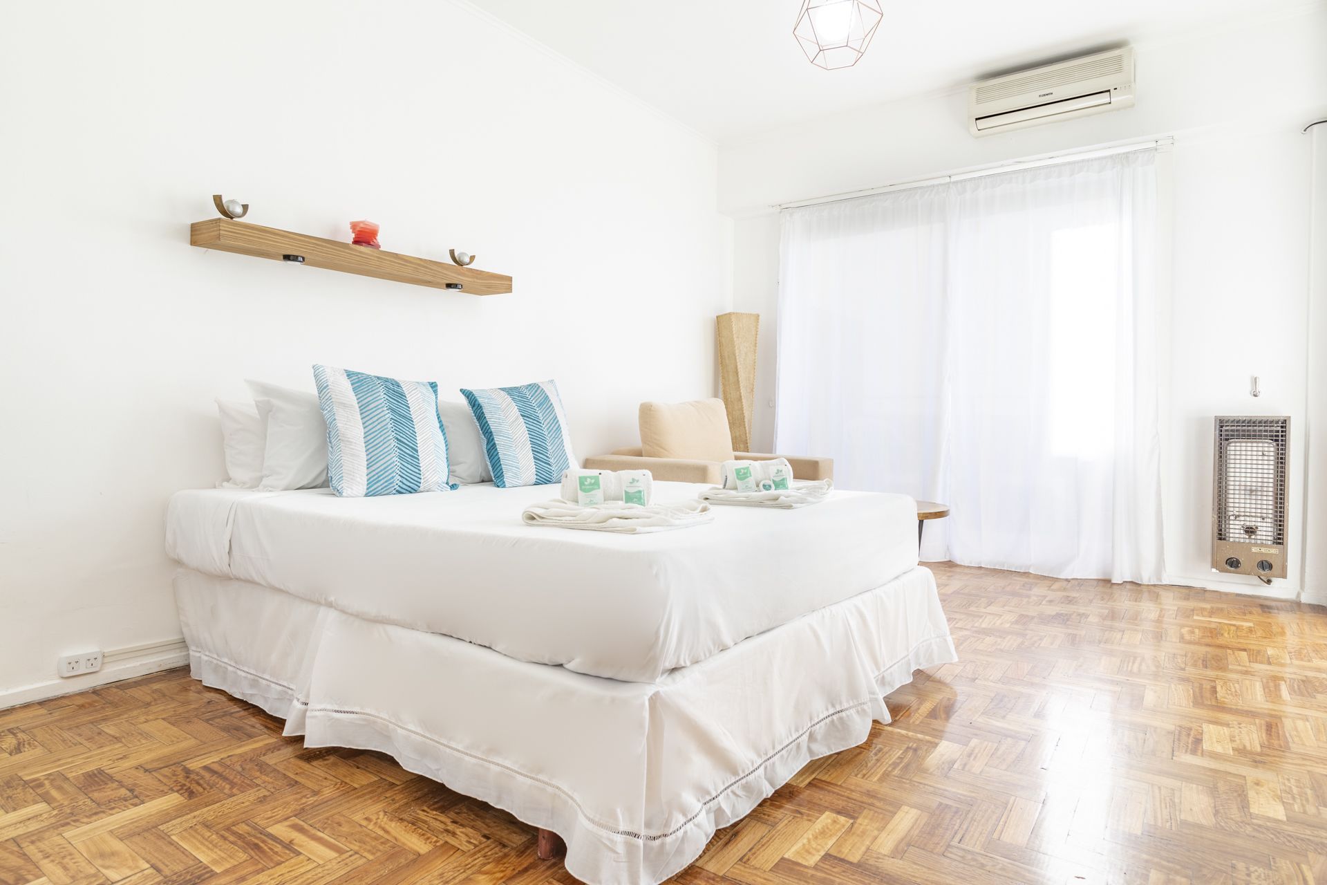 Ein Schlafzimmer mit Holzboden, einem Bett mit weißer Bettwäsche, einem Nachttisch, einer Wandregal mit Dekoration und einem Fenster mit Vorhängen.