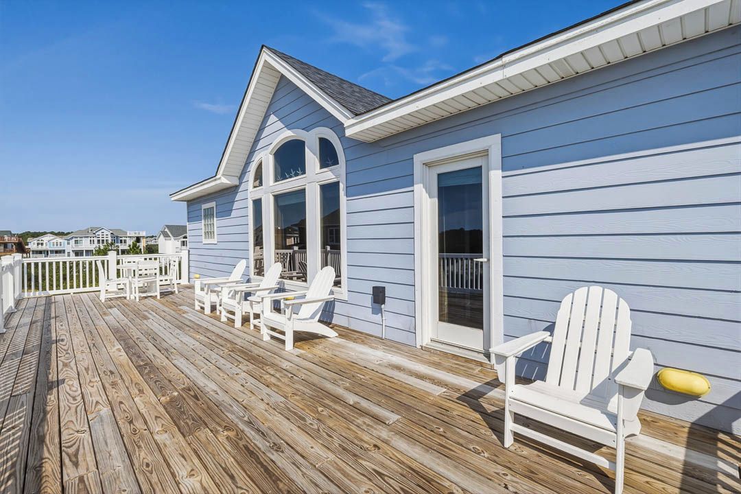 A wooden deck with white Adirondack chairs, a covered area with a white railing, and a light blue building with a gabled roof.
