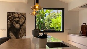 Une cuisine moderne dotée d’un grand îlot central, de luminaires suspendus, d’une fenêtre donnant sur un espace verdoyant et d’une œuvre d’art abstraite accrochée au mur.