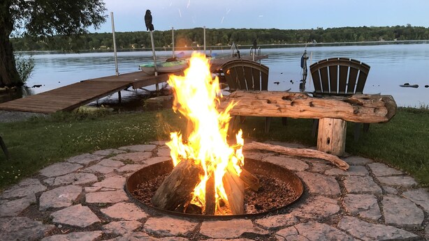 Un focolare in pietra con fuoco acceso, circondato da un pontile di legno e sedie, con vista su un lago.