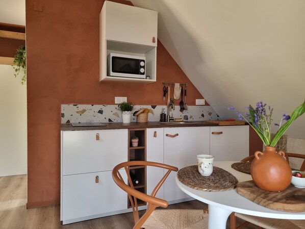 Cocina compacta con armarios blancos, microondas y un lavabo. Hay una mesa redonda con sillas, un florero con flores y un mantel tejido.