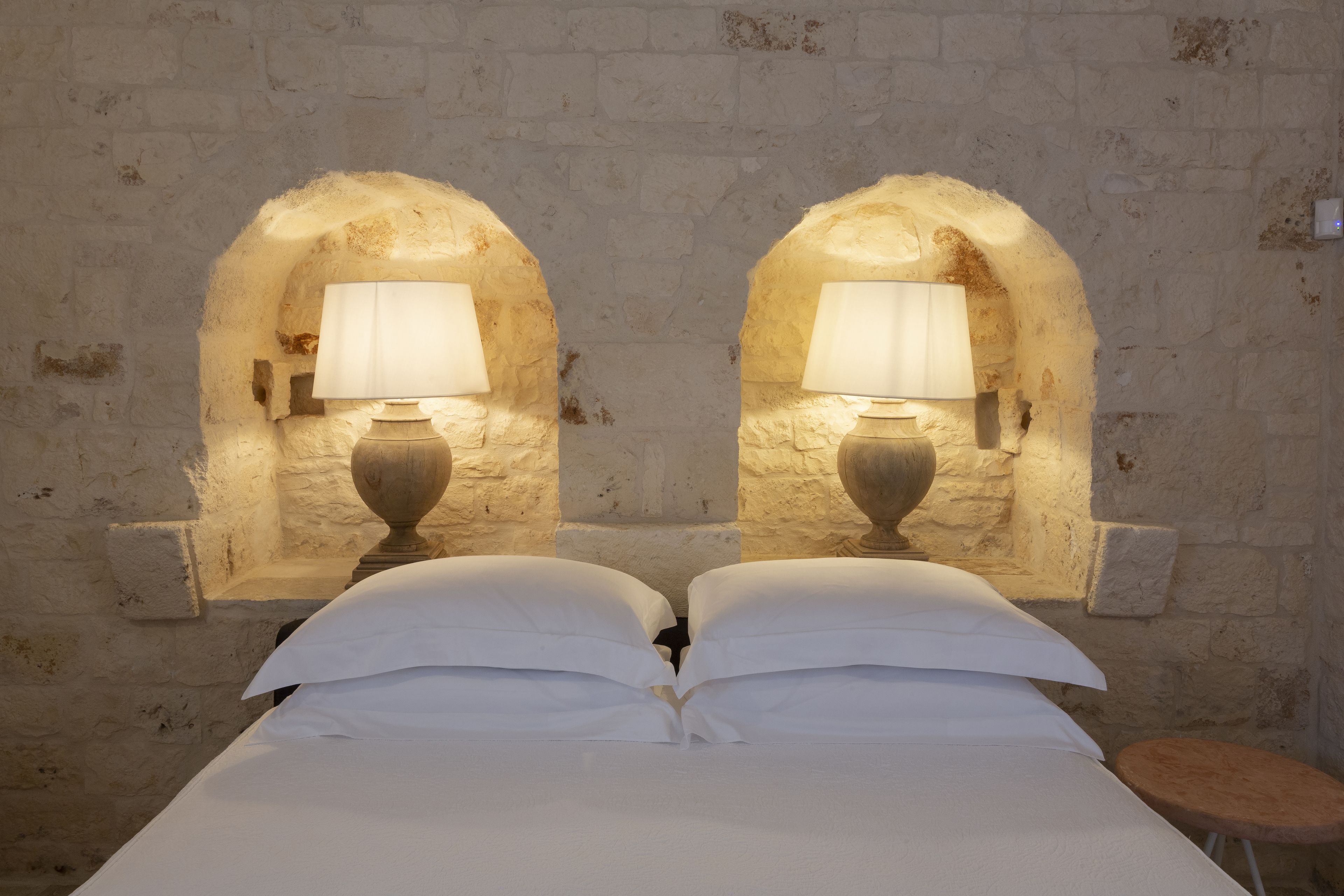 Un letto con lenzuola bianche, due lampade a parete e un muro in mattoni.