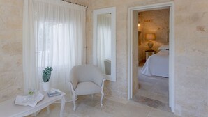 A room with a stone wall, a white chair, a small table with a plant, and a bed in the adjacent room. - Dimora Graziana (Locorotondo)