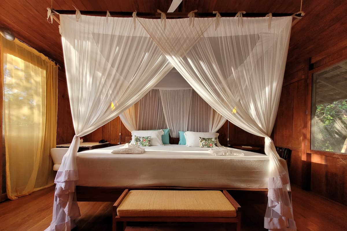 A bedroom with a canopy bed, wooden flooring, and a bench.