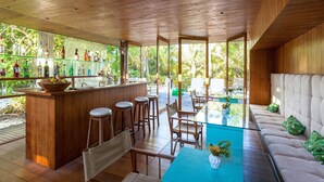 A wooden bar area with stools, a seating area with a sofa and chairs, and a glass table. - Hotel Vila Naia (Prado)