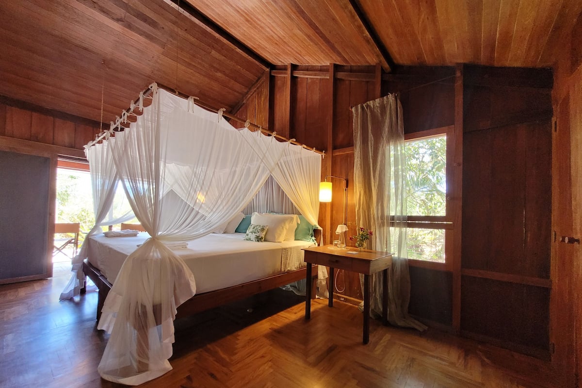 A wooden room with a bed under a canopy, a bedside table, and a window with curtains.