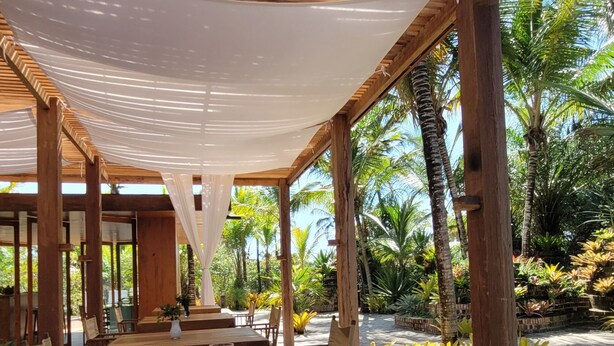 An outdoor dining area with wooden tables and chairs, surrounded by lush greenery and palm trees. - Hotel Vila Naia (Prado)