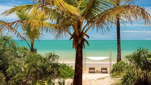 A beach with a palm tree, a white canopy, and two wooden chairs. - Hotel Vila Naia (Prado)
