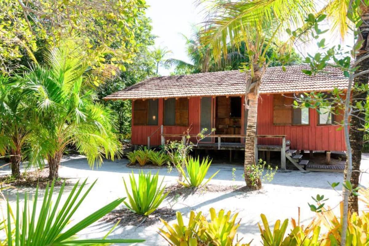A red wooden house with a tiled roof, surrounded by tropical vegetation and a sandy ground.