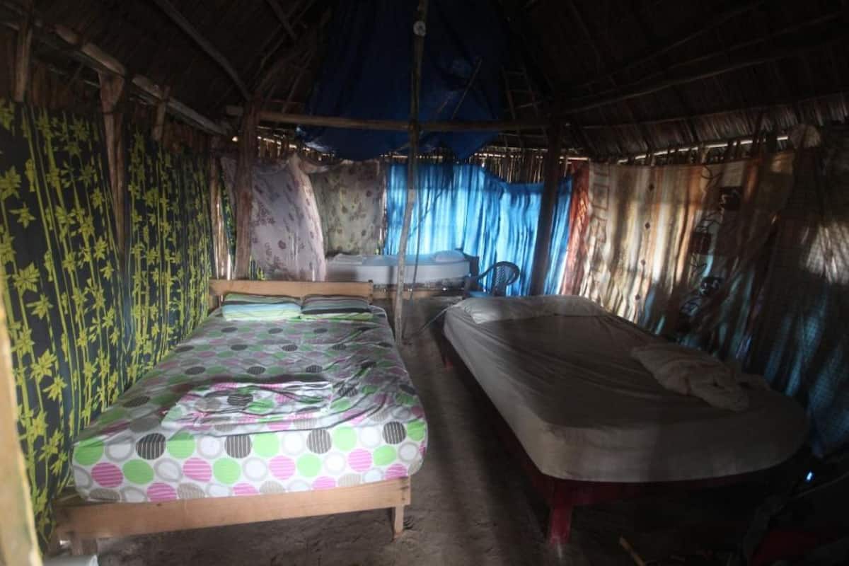 A single bed with a colorful bedspread, a mattress on the floor, and a chair in a rustic room with bamboo walls and a thatched roof.