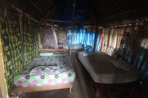 Una cama individual con una colcha colorida, un colchón en el suelo y una silla en una habitación rústica con paredes de bambú y techo de paja.